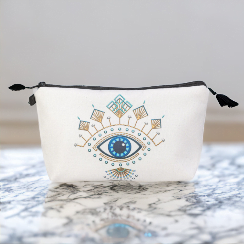 The Roula bag, a white cosmetic bag with artistic blue and gold evil eye pattern on a marble table.