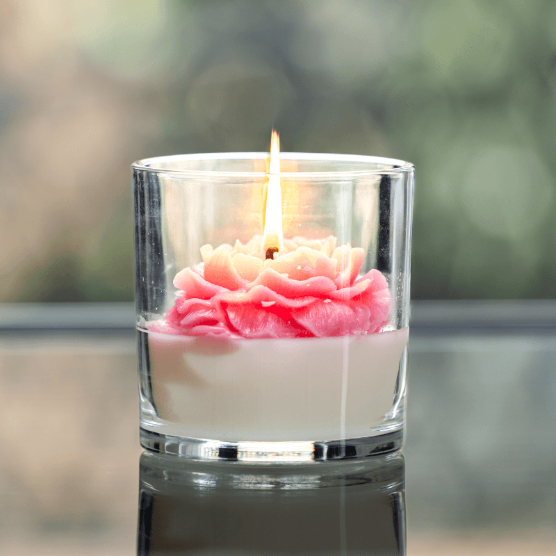 A lit rose candle in a glass jar features a pink and white flower-shaped wax design.